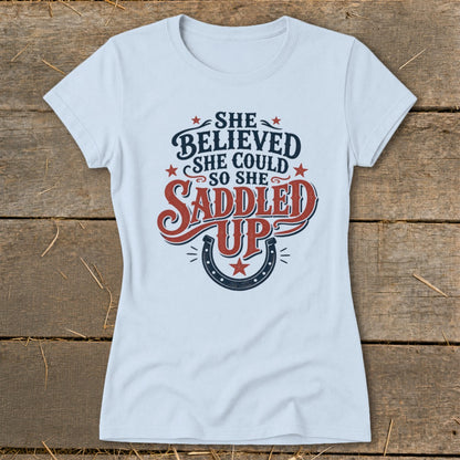 Light blue t-shirt with "She believed she could so she saddled up" text and horseshoe design.