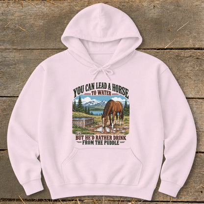 A light pink hooded sweatshirt features a graphic of a horse drinking from a puddle with mountain scenery.