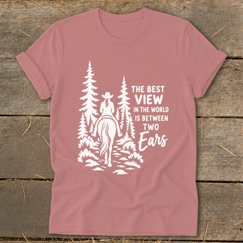 Mauve t-shirt featuring a white graphic of a cowgirl on horseback amid pine trees, with text reading The best view in the world is between two ears.