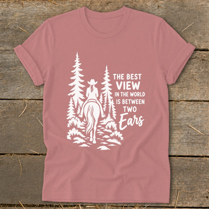 Mauve t-shirt featuring a white graphic of a cowgirl on horseback amid pine trees, with text reading The best view in the world is between two ears.
