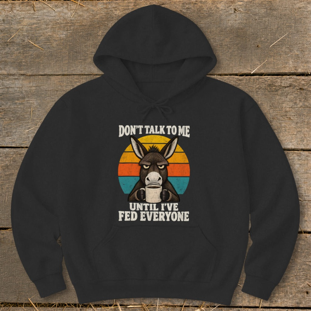 A black hooded sweatshirt featuring a grumpy donkey holding a coffee mug with sunset-colored stripes behind it and text reading "Don't Talk to Me Until I've Fed Everyone".