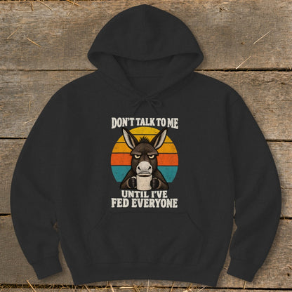 A black hooded sweatshirt featuring a grumpy donkey holding a coffee mug with sunset-colored stripes behind it and text reading "Don't Talk to Me Until I've Fed Everyone".