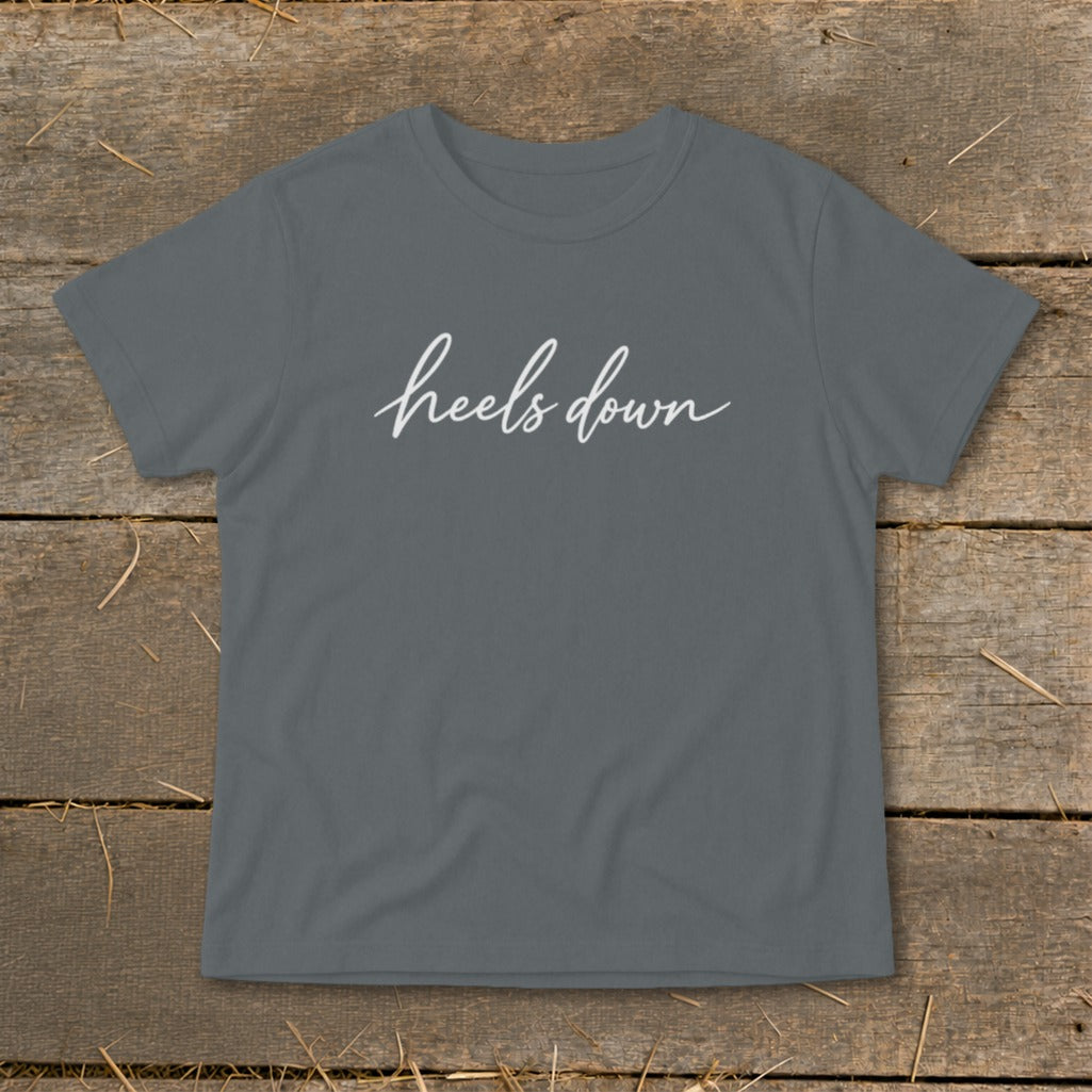 Grey t-shirt with white "heels down" script text on a wooden background.