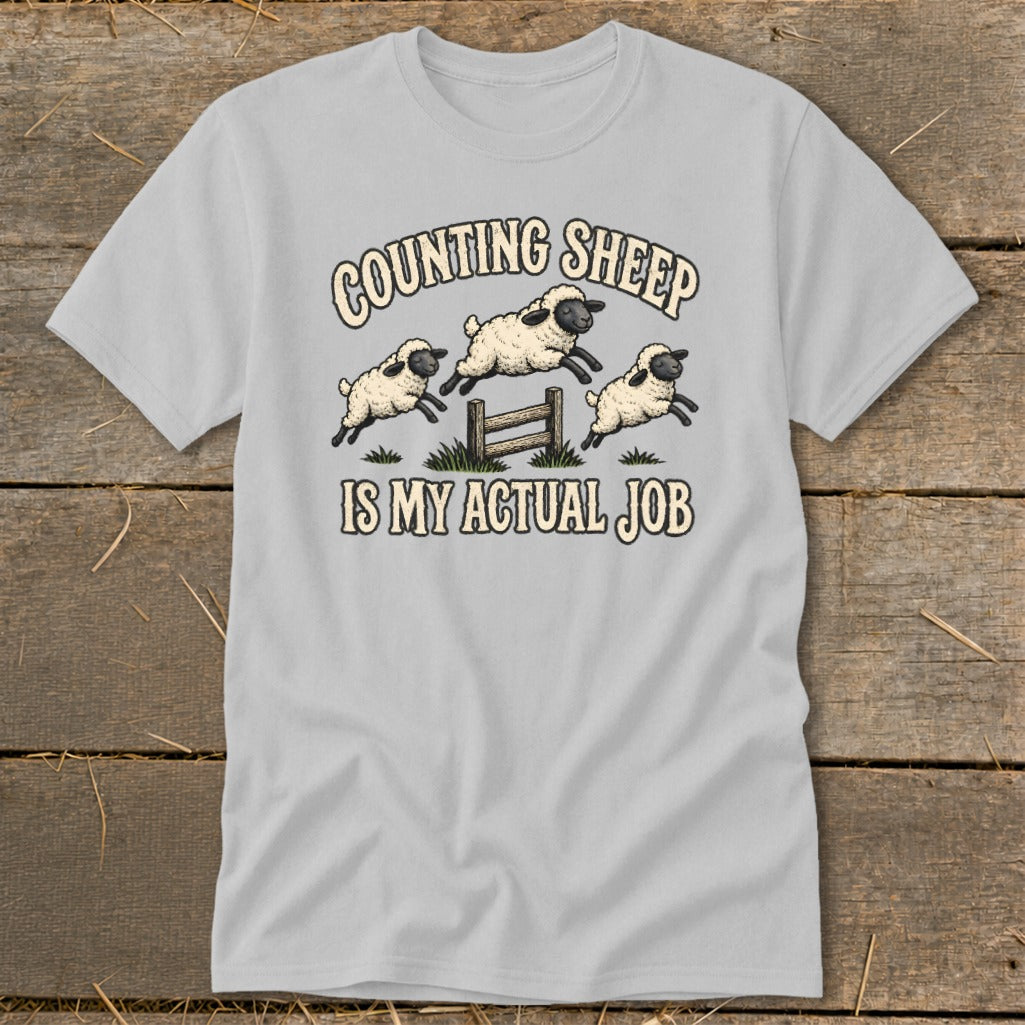 Light gray t-shirt with cartoon sheep jumping over a fence and text "COUNTING SHEEP IS MY ACTUAL JOB".