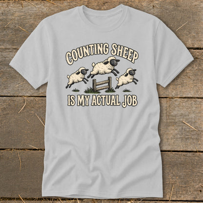Light gray t-shirt with cartoon sheep jumping over a fence and text "COUNTING SHEEP IS MY ACTUAL JOB".