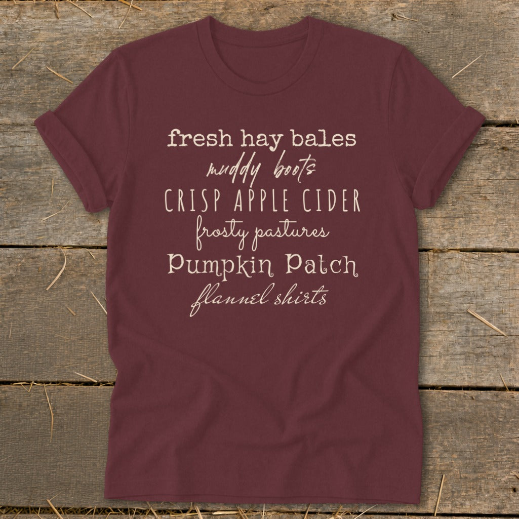 Maroon t-shirt with list: fresh hay bales, muddy boots, crisp apple cider, frosty pastures, pumpkin patch, flannel shirts.