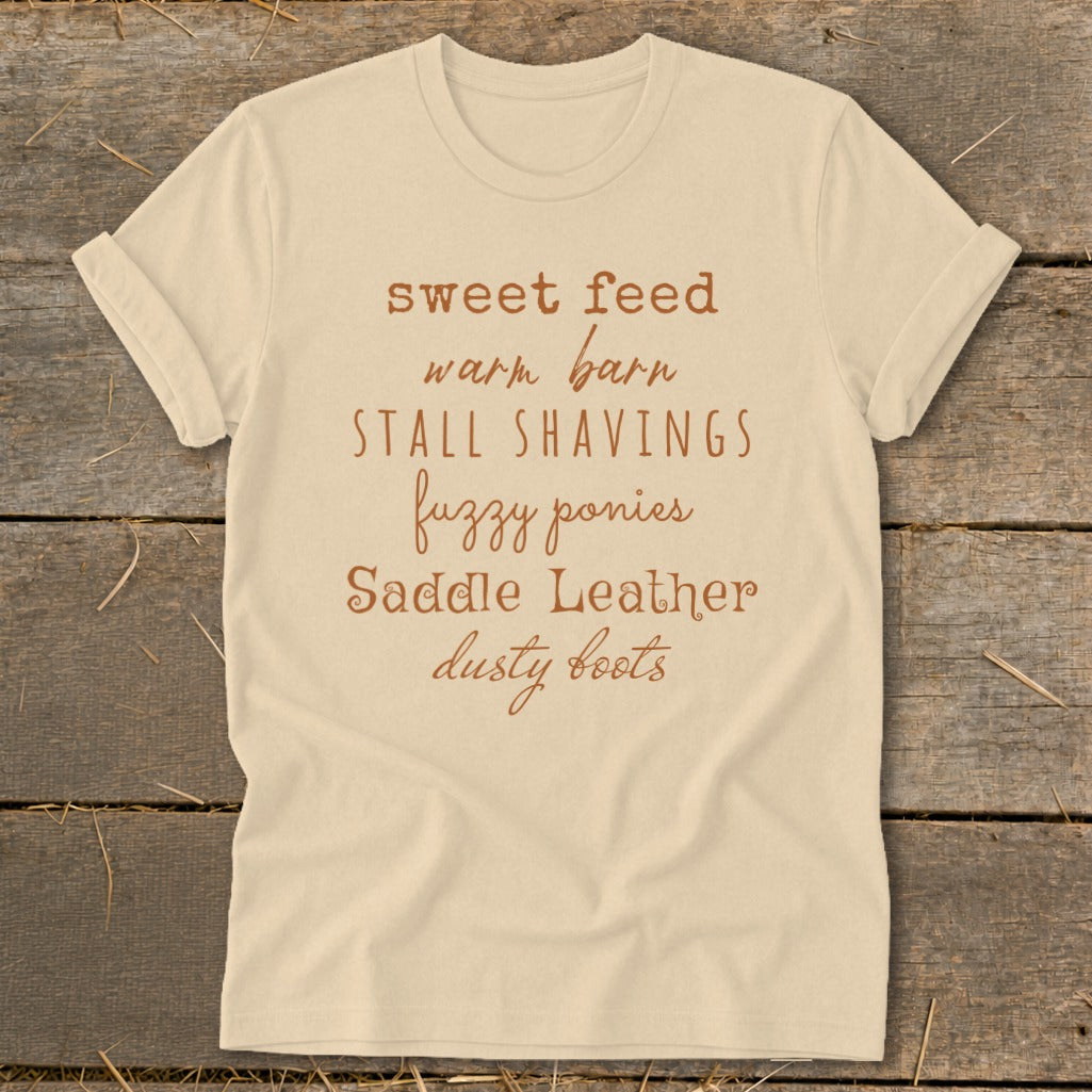 Cream t-shirt with brown text listing "sweet feed, warm barn, stall shavings, fuzzy ponies, saddle leather, dusty boots."