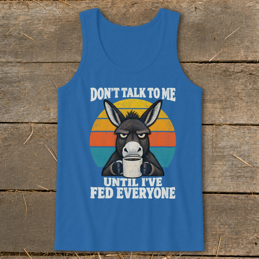Blue tank top featuring a grumpy donkey drinking coffee in front of a sunset, with text saying, "DON'T TALK TO ME UNTIL I'VE FED EVERYONE."