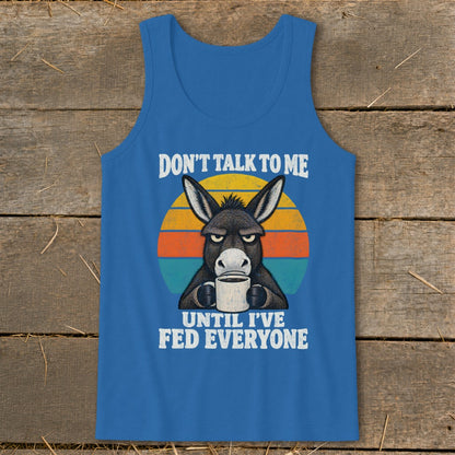 Blue tank top featuring a grumpy donkey drinking coffee in front of a sunset, with text saying, "DON'T TALK TO ME UNTIL I'VE FED EVERYONE."