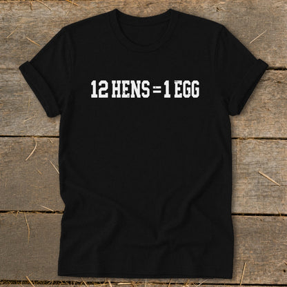 Black short-sleeve t-shirt with rolled cuffs featuring the white text 12 HENS = 1 EGG laid flat on wood.
