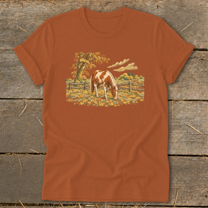 Brown and white horse grazing in autumn field with wooden fence and colorful trees.
