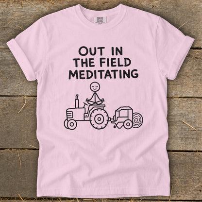 Pink t-shirt with "Out in the Field Meditating" text and stick figure meditating on tractor.