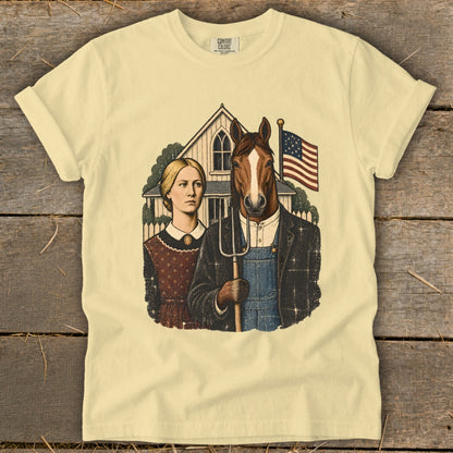 A light yellow t-shirt with a vintage graphic featuring a woman and a horse-headed farmer holding a pitchfork in front of a house and an American flag.
