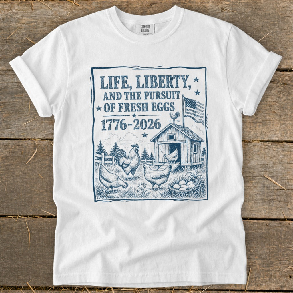 White t-shirt with a blue graphic of chickens, a coop, and text: "Life, Liberty, and the Pursuit of Fresh Eggs 1776-2026."