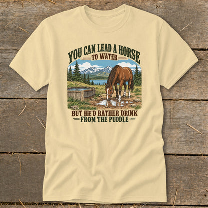 A light yellow t-shirt featuring a graphic of a horse drinking from a puddle with humorous text.