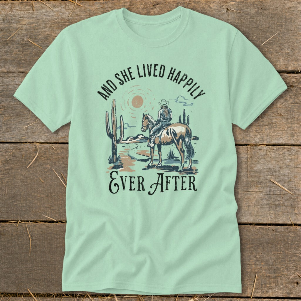 Light green t-shirt with graphic of a woman riding a horse in a desert landscape with cacti and the text "AND SHE LIVED HAPPILY EVER AFTER".