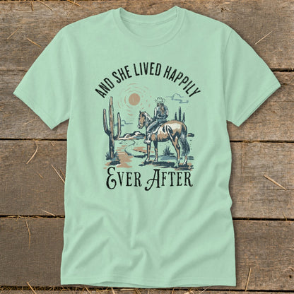 Light green t-shirt with graphic of a woman riding a horse in a desert landscape with cacti and the text "AND SHE LIVED HAPPILY EVER AFTER".