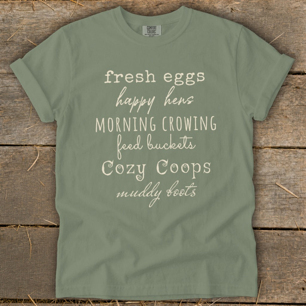 Sage t-shirt with "fresh eggs happy hens morning crowing feed buckets cozy coops muddy boots" text.