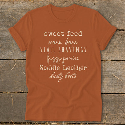Orange t-shirt with white text: "sweet feed warm barn STALL SHAVINGS fuzzy ponies Saddle Leather dusty boots".