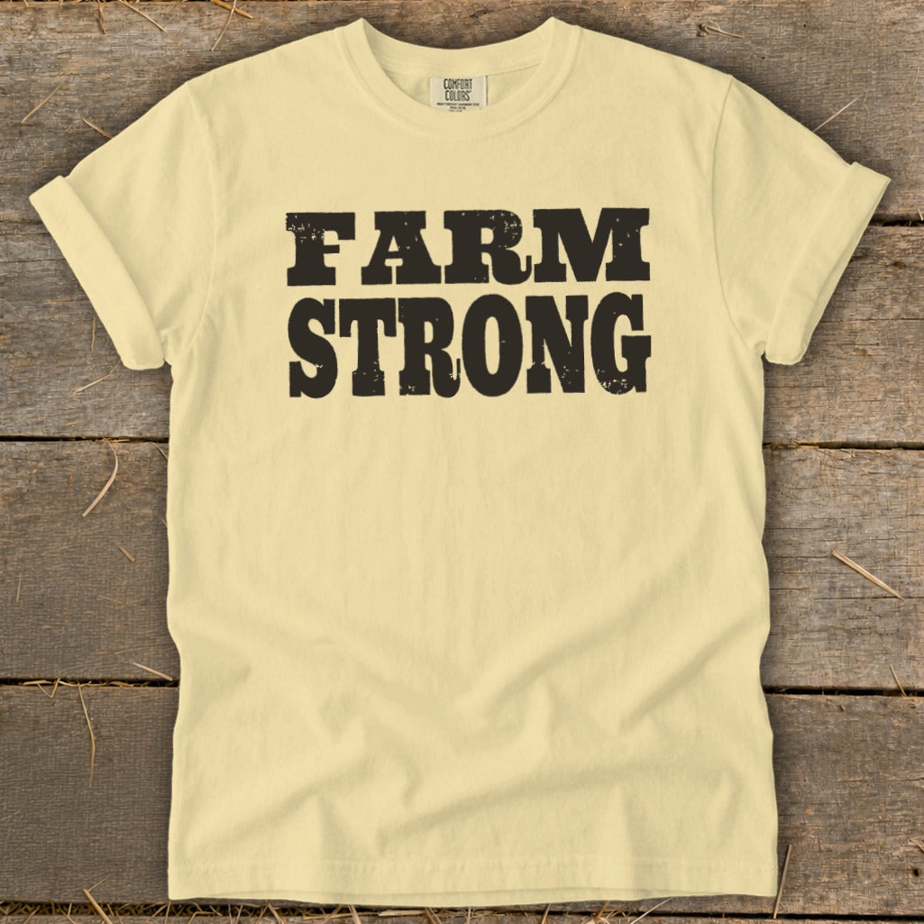 A light yellow t-shirt with rolled sleeves laid flat on a rustic wooden background, featuring bold black distressed text that reads FARM STRONG across the chest.