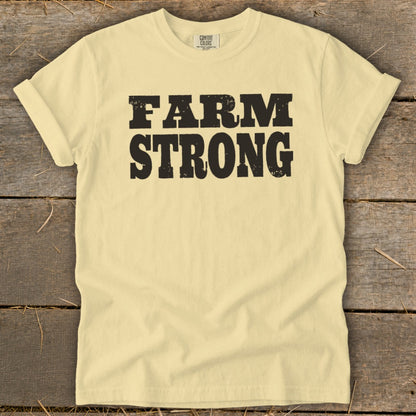 A light yellow t-shirt with rolled sleeves laid flat on a rustic wooden background, featuring bold black distressed text that reads FARM STRONG across the chest.