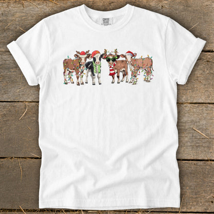Five cartoon cows wear Santa hats and Christmas lights on a white t-shirt.