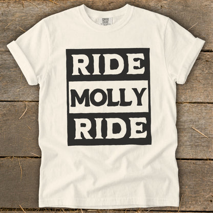 A cream-colored t-shirt on a wooden surface with the text "RIDE MOLLY RIDE" in bold, block letters.