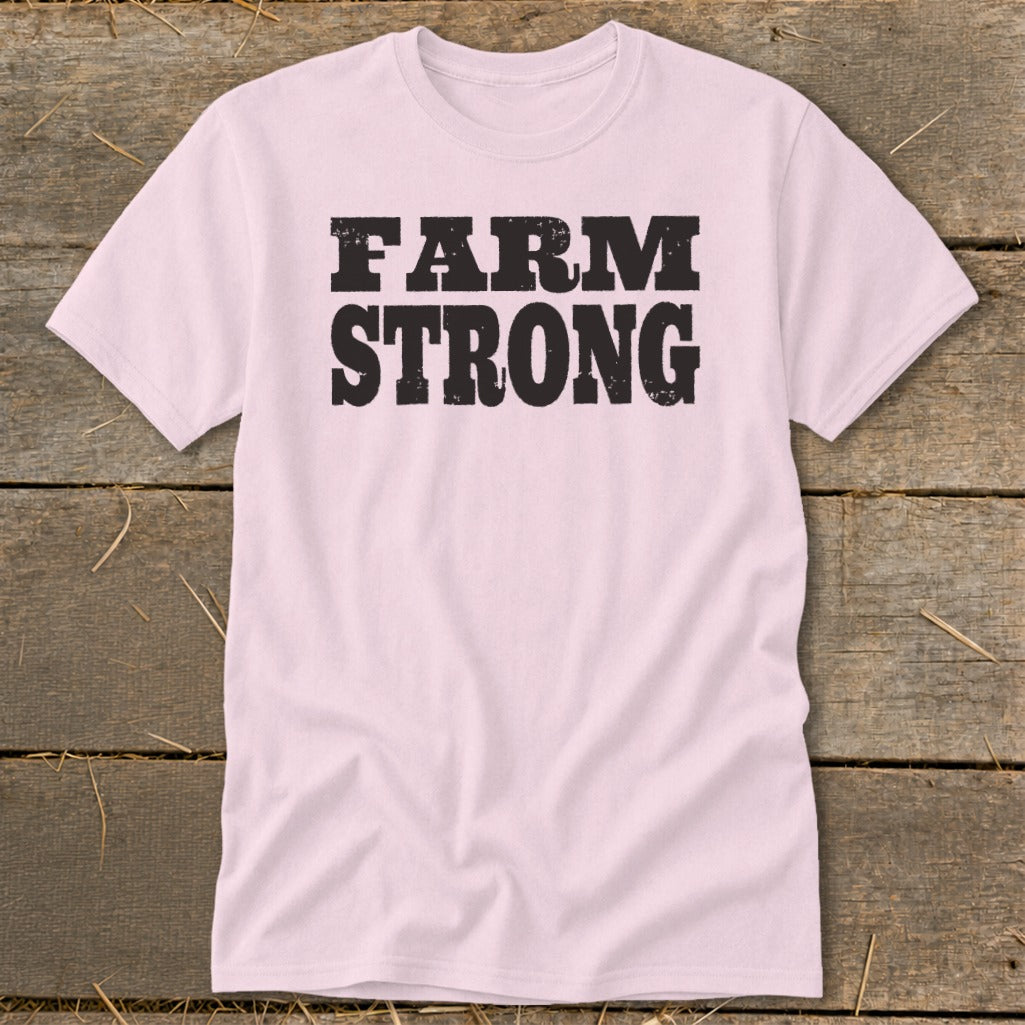 A light pink t-shirt with black distressed text reading FARM STRONG on the chest, lying flat on a wooden surface.
