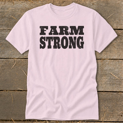 A light pink t-shirt with black distressed text reading FARM STRONG on the chest, lying flat on a wooden surface.