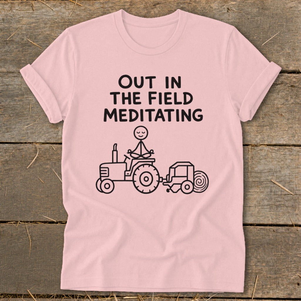 Pink t-shirt with black text "OUT IN THE FIELD MEDITATING" and stick figure meditating on a tractor.