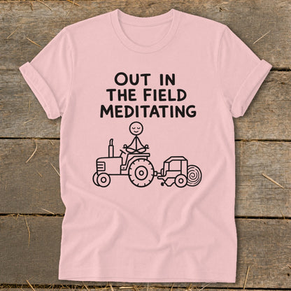 Pink t-shirt with black text "OUT IN THE FIELD MEDITATING" and stick figure meditating on a tractor.