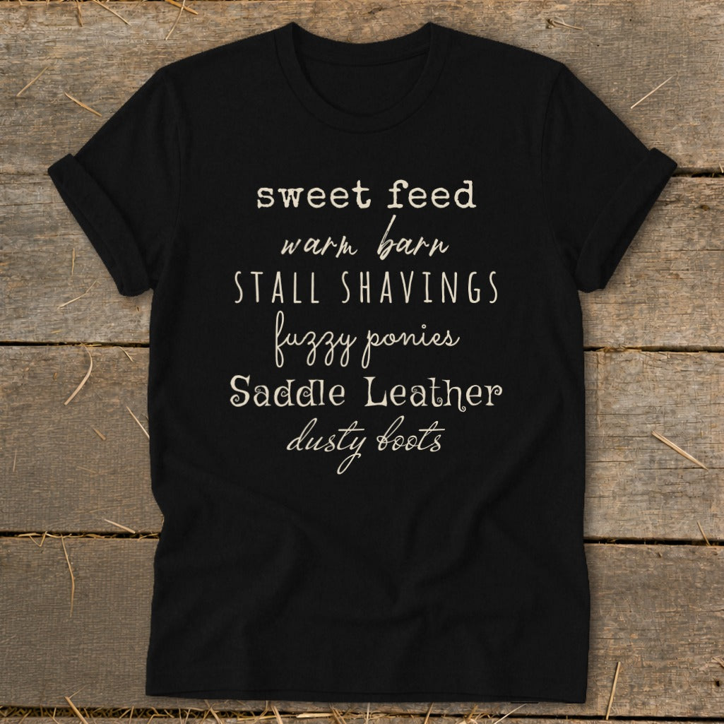 Black t-shirt with text: sweet feed, warm barn, stall shavings, fuzzy ponies, saddle leather, dusty boots.