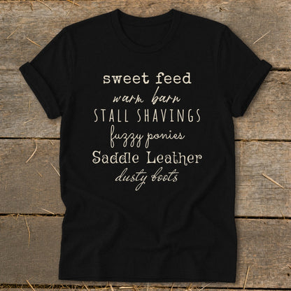 Black t-shirt with text: sweet feed, warm barn, stall shavings, fuzzy ponies, saddle leather, dusty boots.