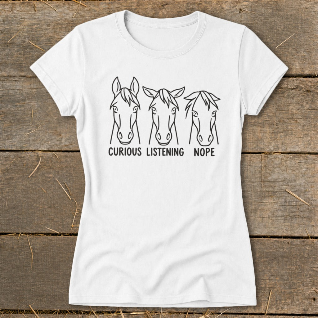 White t-shirt featuring three horse head sketches with text reading curious listening nope displayed on a wooden surface.