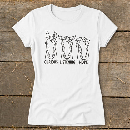 White t-shirt featuring three horse head sketches with text reading curious listening nope displayed on a wooden surface.