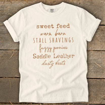 White t-shirt with text: sweet feed, warm barn, stall shavings, fuzzy ponies, saddle leather, dusty boots.