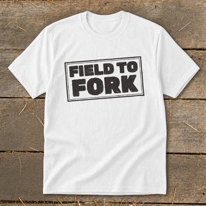 A white crewneck t-shirt featuring a black rectangular stamp design with the text FIELD TO FORK centered on a rustic wooden background.