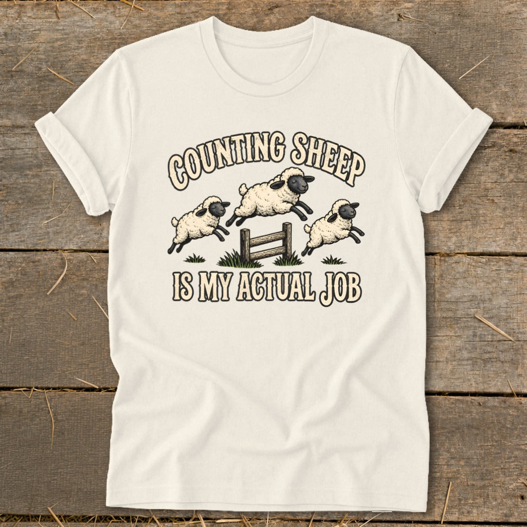 Cream t-shirt with illustration of three sheep jumping over a fence, text "COUNTING SHEEP IS MY ACTUAL JOB".