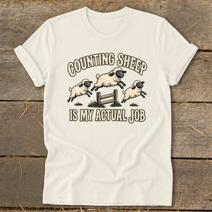 Cream t-shirt with illustration of three sheep jumping over a fence, text "COUNTING SHEEP IS MY ACTUAL JOB".