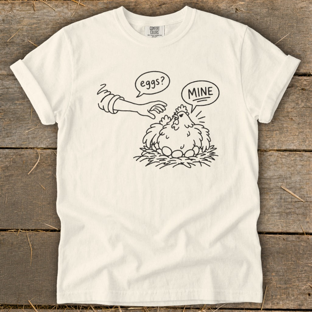 Line drawing on shirt: a chicken on a nest with eggs, a hand reaches in saying "eggs?", chicken says "MINE".