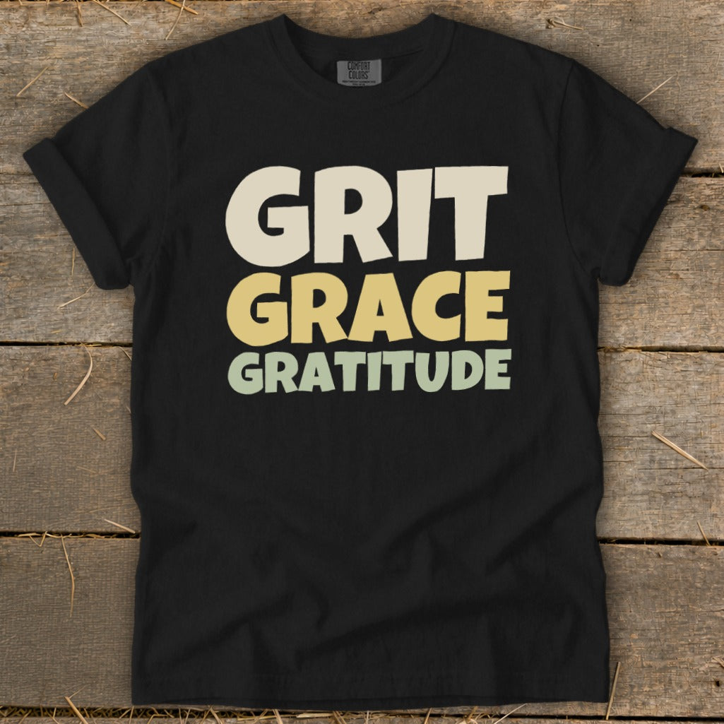Black t-shirt featuring stacked retro text that reads Grit Grace Gratitude on a rustic wooden background.