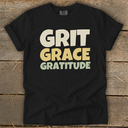 Black t-shirt featuring stacked retro text that reads Grit Grace Gratitude on a rustic wooden background.