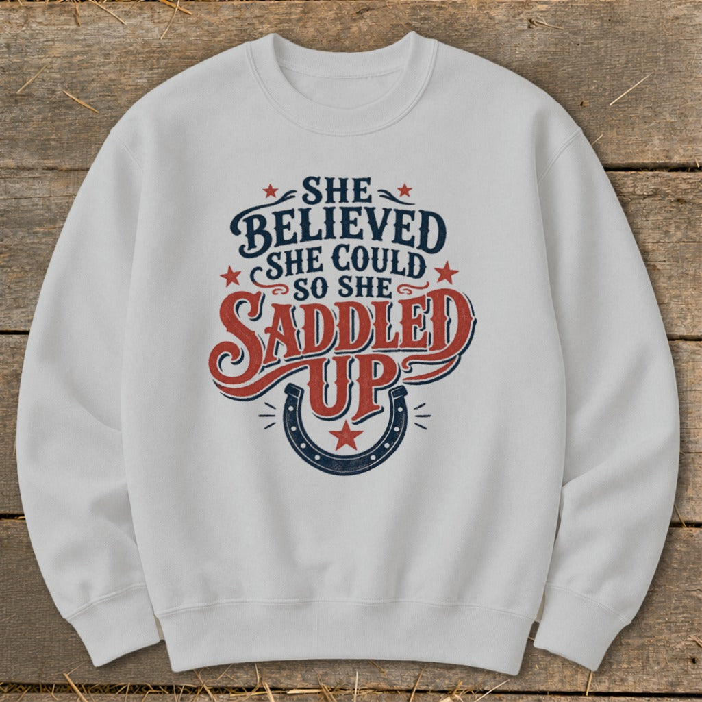 White sweatshirt with "She believed she could so she saddled up" text and horseshoe graphic.