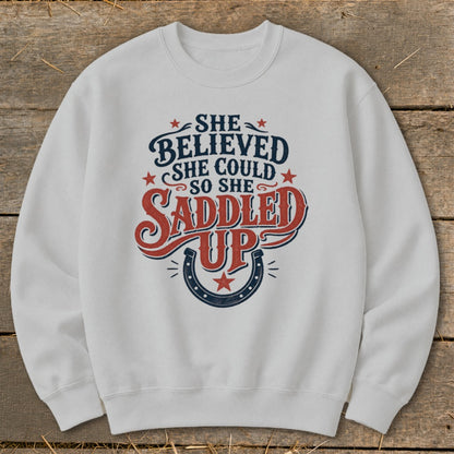 White sweatshirt with "She believed she could so she saddled up" text and horseshoe graphic.