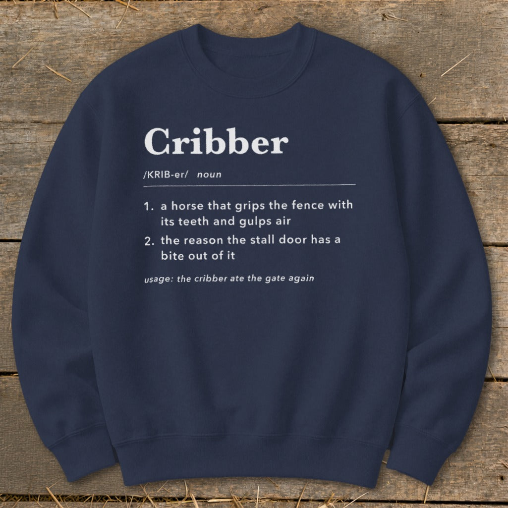 Navy blue crewneck sweatshirt featuring white text defining the noun Cribber laid flat on wood.