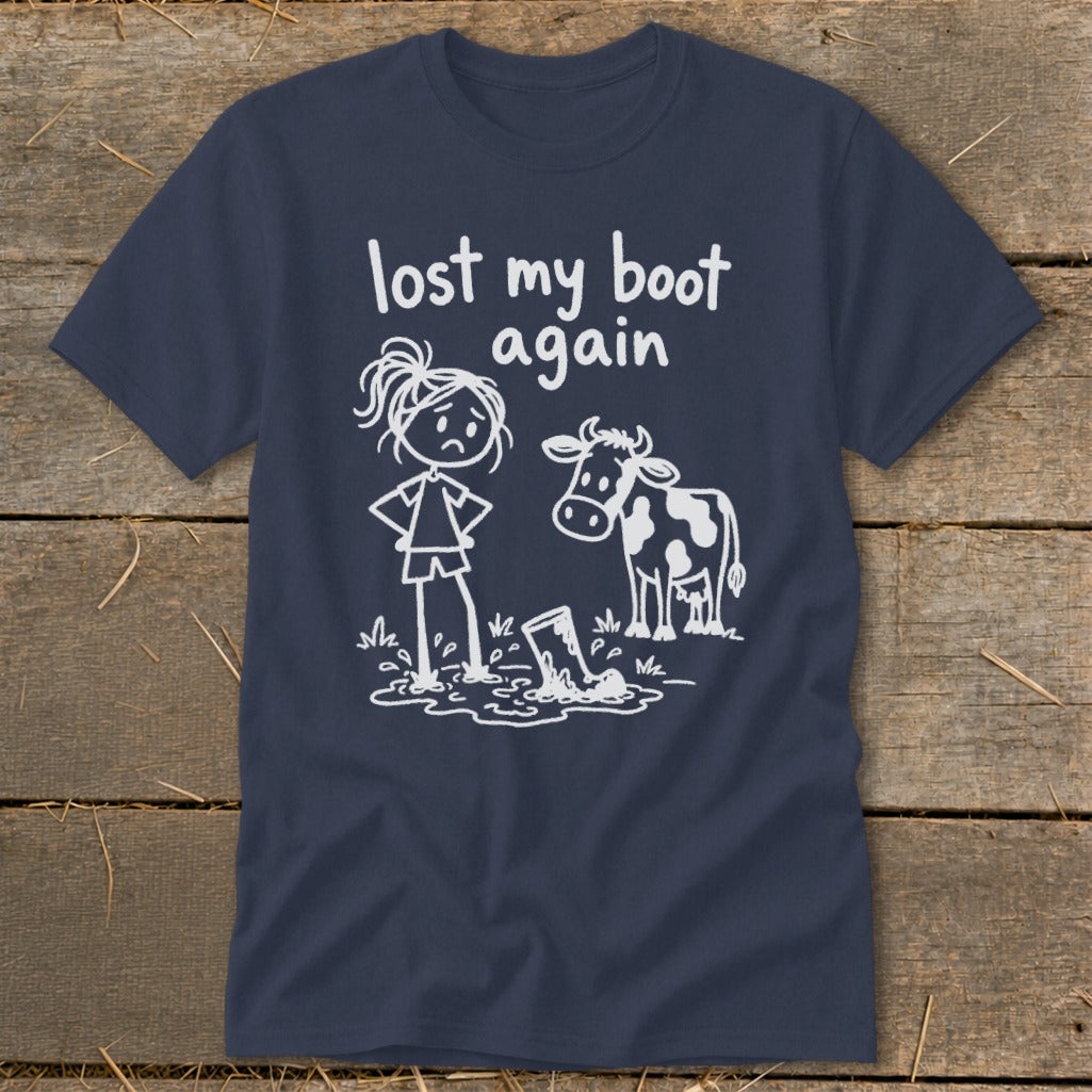 A dark blue t-shirt with a white graphic of a frustrated stick-figure cowgirl and a cow standing near a boot stuck in mud, accompanied by the text "lost my boot again."