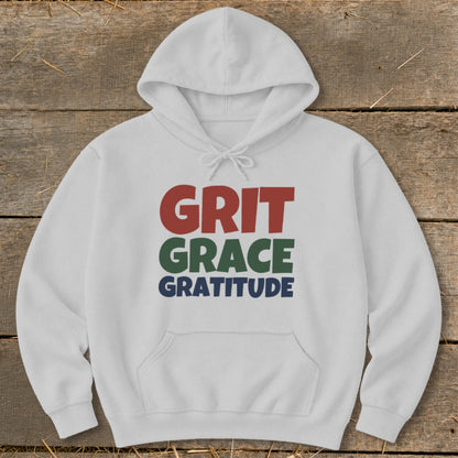 White hooded sweatshirt laid flat on a rustic wooden background with multicolored text reading GRIT GRACE GRATITUDE centered on the chest.