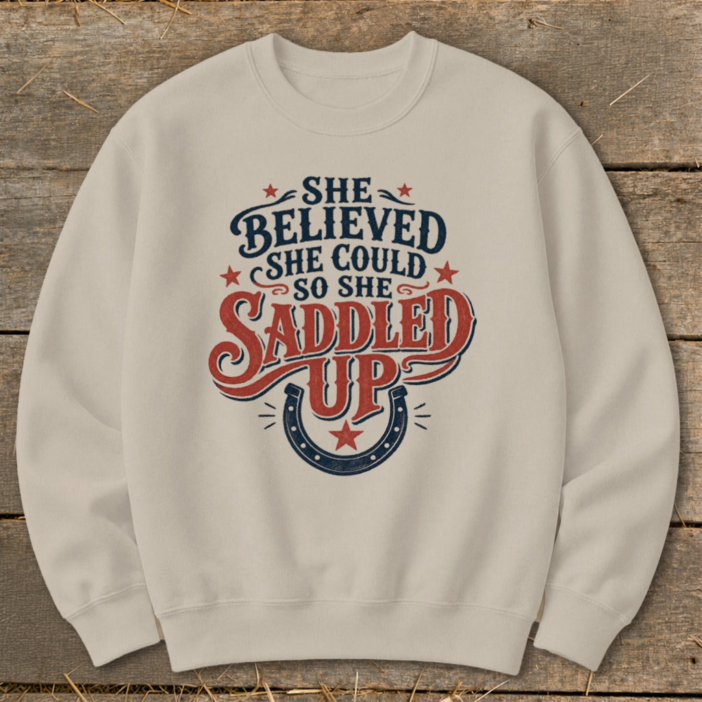 Beige sweatshirt with "She Believed She Could So She Saddled Up" text and horseshoe graphic.