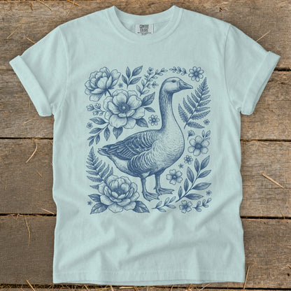 Light blue t-shirt with a navy blue sketch of a goose surrounded by flowers and ferns.
