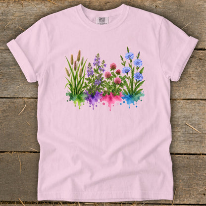 A light pink t-shirt featuring a watercolor botanical print of wildflowers over colorful paint splatters.